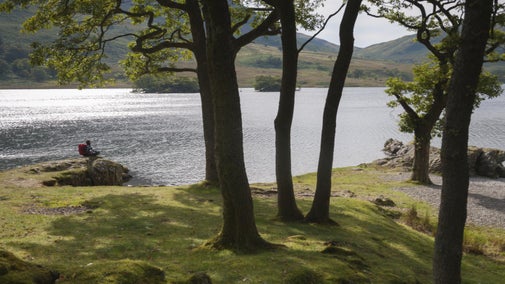 Backpacker sitting on ground looking over lake with trees in foreground and large hills beyond the water